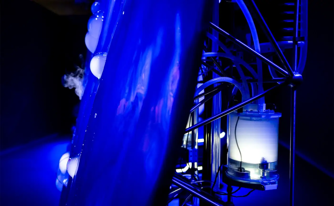 Close-up of the Symbiosis mechanism: a lit fluid reservoir and tubing on a steel frame beside the blue sculpture